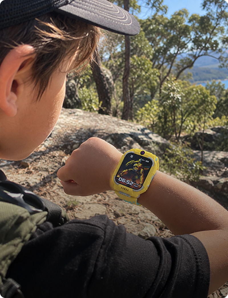 A child outdoors wearing a bright yellow kids' smartwatch with a colorful display, shown in a natural rocky forest setting.