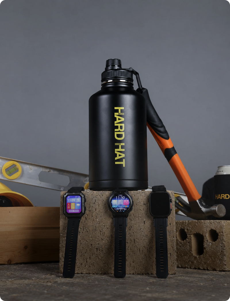 A matte black Hard Hat branded water bottle with a carry loop stands on a concrete block, with three smartwatches in front of it and construction tools in the background against a gray backdrop.